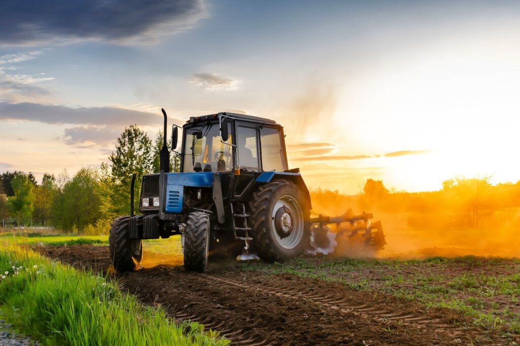 Modern blue tractor machinery plowing agricultural field meadow at farm at spring during sunset